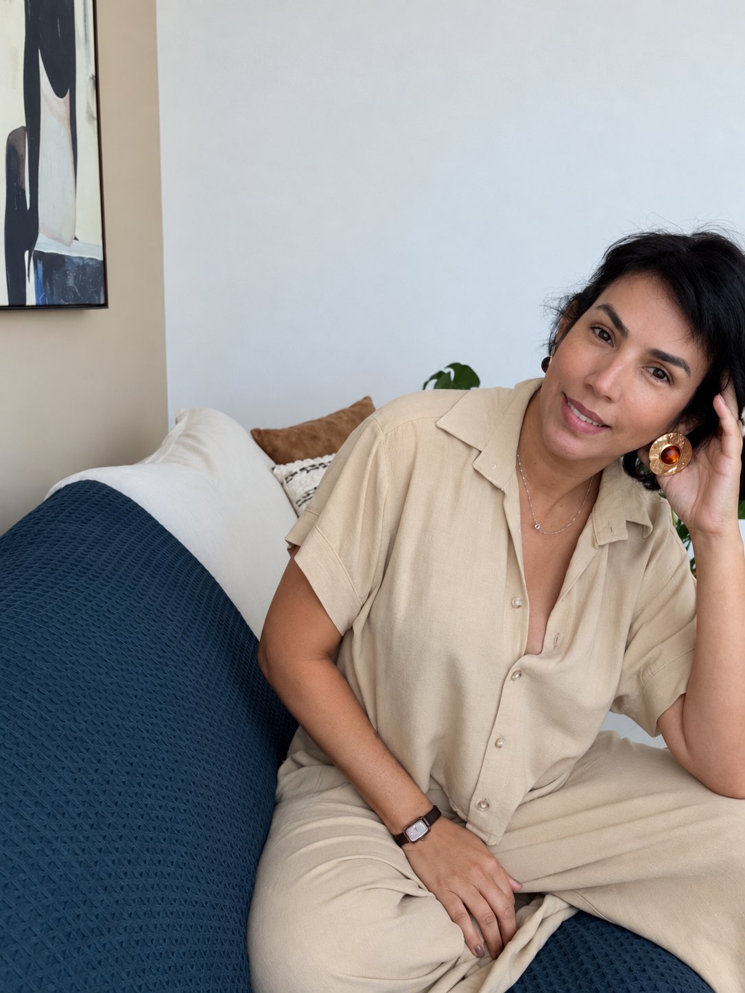 Priscila Honorato sentada em um sofá com manta azul, vestindo um conjunto bege